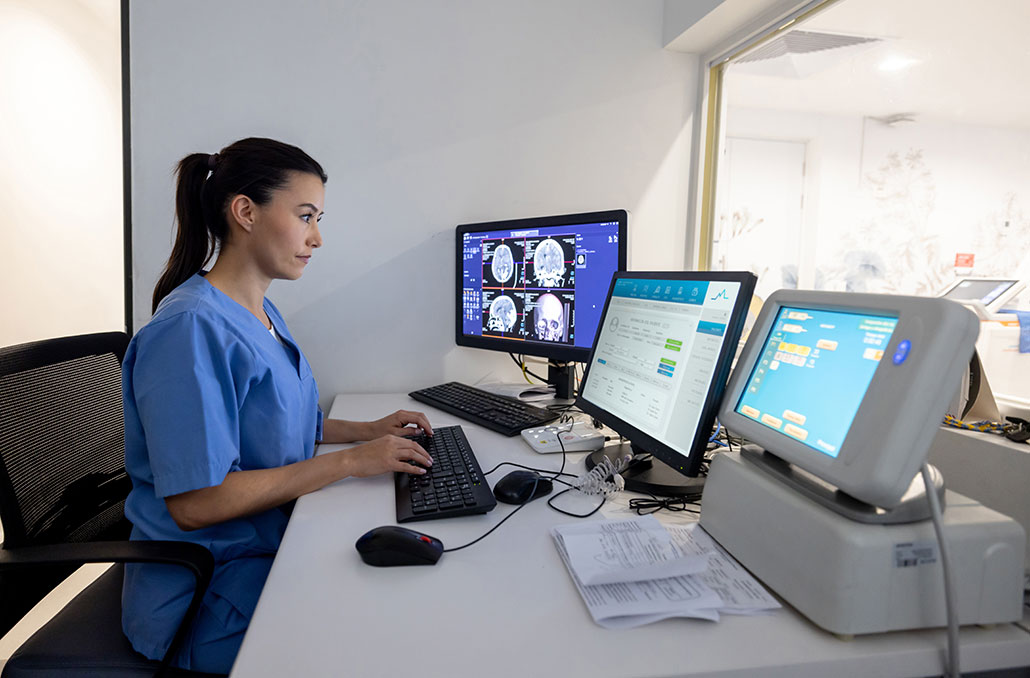 A woman sitting in front of a computer and a medical device, screens show an image of a person's skull